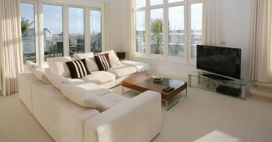 Amplio salón con sofá en tonos claros, mesa de centro de madera y grandes ventanales que dejan entrar la luz natural y ofrecen vistas al exterior.