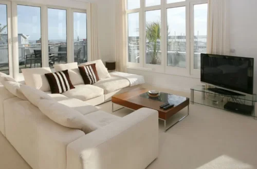 Amplio salón con sofá en tonos claros, mesa de centro de madera y grandes ventanales que dejan entrar la luz natural y ofrecen vistas al exterior.