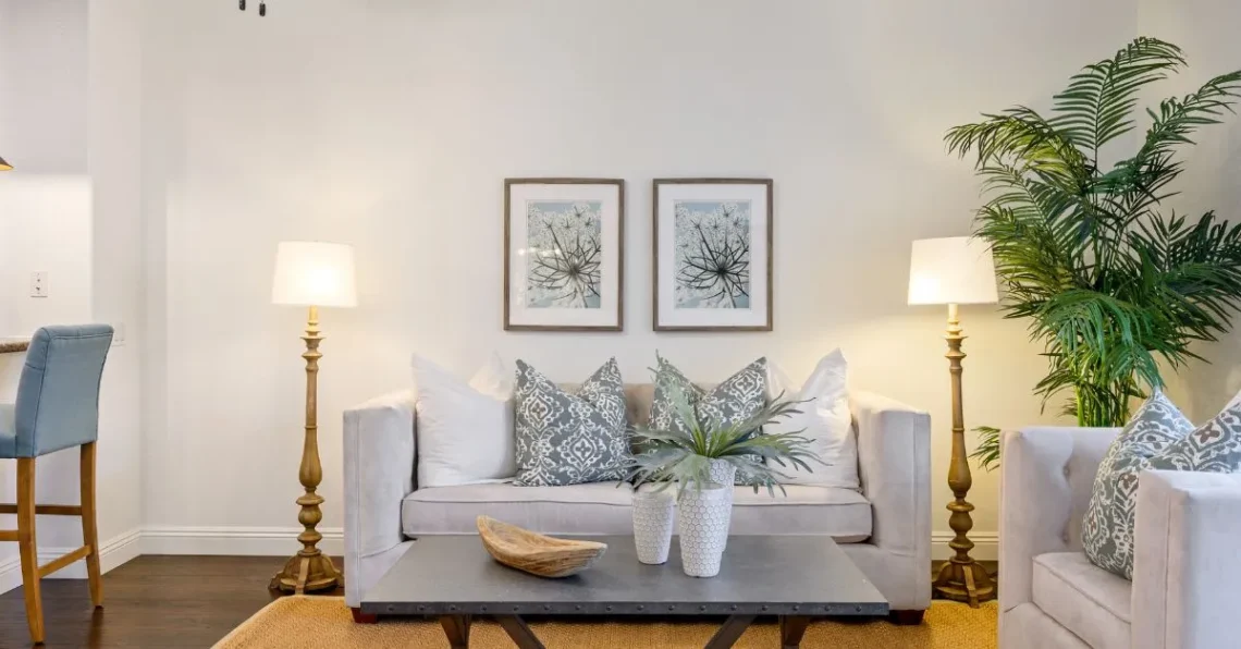 Salón con sofá central, cojines en tonos neutros y estampados suaves, iluminación simétrica con lámparas de pie y cuadros decorativos que crean un ambiente equilibrado, ordenado y acogedor.