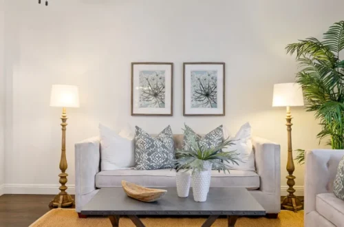 Salón con sofá central, cojines en tonos neutros y estampados suaves, iluminación simétrica con lámparas de pie y cuadros decorativos que crean un ambiente equilibrado, ordenado y acogedor.