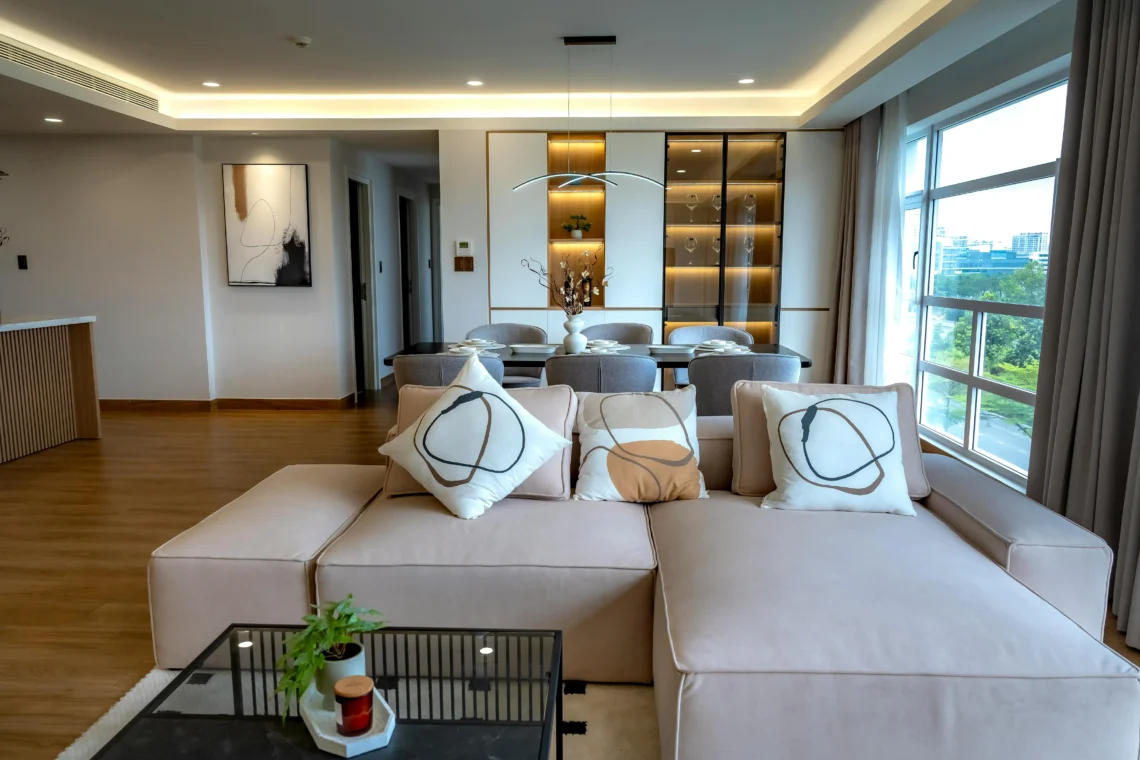 Sala de estar moderna con un sofá modular beige y cojines decorativos abstractos, conectada a un comedor con sillas grises y una vitrina iluminada al fondo. Grandes ventanales muestran una vista urbana luminosa.