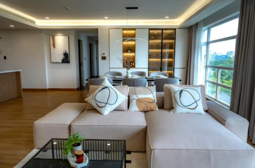 Sala de estar moderna con un sofá modular beige y cojines decorativos abstractos, conectada a un comedor con sillas grises y una vitrina iluminada al fondo. Grandes ventanales muestran una vista urbana luminosa.