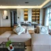 Sala de estar moderna con un sofá modular beige y cojines decorativos abstractos, conectada a un comedor con sillas grises y una vitrina iluminada al fondo. Grandes ventanales muestran una vista urbana luminosa.